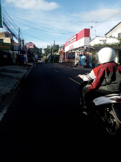 The image depicts a street scene in the city of Khouathineung, Laos, observed from behind a person operating a motor scooter. The road surface is asphalt, with a faded yellow line visible on the right side, indicating a lane division or shoulder.
In the immediate foreground, the back of a person wearing a white full-face helmet is visible. This individual is seated on a white and black scooter, distinguished by a white rear fender and exposed mechanical components. They are wearing a red and grey jacket, patterned trousers, and black Adidas slide sandals with white stripes. Their hands grip the handlebars, though specific controls are not clear.
The street extends into the distance, appearing to be a two-lane road or a single lane with a wide shoulder, accommodating traffic in both directions. Several vehicles, primarily motor scooters and at least one white car, are visible further down the road, receding from the viewer.
On the left side of the road, a series of commercial buildings and residences are present. A prominent sign for "Alfamart," a convenience store, is clearly visible. Below it, utility poles are laden with numerous overhead electrical and communication wires. Another building features a partially obscured sign. The pavement on the left appears uneven and partially covered with dirt or debris.
On the right side of the road, a large building with a distinctive red and white facade dominates, displaying a sign that reads "TOKO JAYA" and "Triple Tone." This structure also has an awning and appears to be a commercial establishment, possibly a hardware or general goods store. Below this building, a patterned barrier or fence is visible. More overhead wires are present on this side of the street.
The background features distant residential structures, green foliage, and trees. The sky is bright blue with scattered white clouds, indicating clear weather. Sunlight illuminates the scene, creating noticeable shadows on the left side of the road and foreground, suggesting either morning or late afternoon.