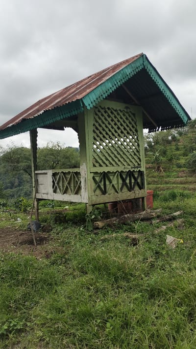 This image depicts a weathered, abandoned wooden gazebo situated on a grassy incline in Payatumpi Baru, Indonesia. The gazebo has a rusted corrugated metal roof with decorative green trim. Its walls are constructed from light green lattice work and white wooden planks, some with an 'X' pattern. A broken shovel leans against one of the supporting posts. The surrounding environment features lush green grass, scattered debris, trees, and terraced fields in the background, indicating a rural or semi-rural setting. The sky is overcast, suggesting a cloudy or impending rainy day. There are no people or animals visible in the scene.