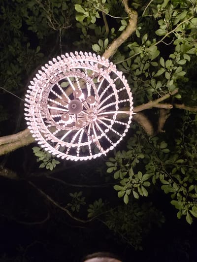 An ornate, illuminated chandelier hangs from a tree branch at night in Bettembourg, Luxembourg. The chandelier is made of white metal and appears to be decorated with numerous small, reflective elements, possibly crystals, creating a circular, spoked design with a central light fixture. The surrounding environment is dark, with the tree's leaves visible in the upper and right portions of the image, illuminated by the chandelier's warm light. The tree trunk is visible to the left of the chandelier, with its bark texture apparent.