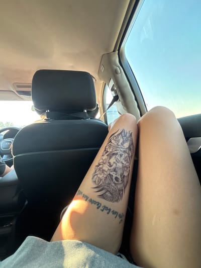 The image shows the interior of a car from the perspective of a passenger. The focus is on the passenger's legs and a tattoo on the left thigh, which depicts a skull surrounded by flowers and a snake. The tattoo has text underneath, partially visible as "love her but leave her will." The passenger is wearing a gray garment, possibly shorts or a skirt. In the background, a headrest and part of a car seat are visible. The steering wheel of the car, with an Audi logo, is also visible on the left side. The sky is visible through the car windows, indicating it is daytime with clear weather. The location is not specified as Mikrevo, Bulgaria, nor is there any information in the image to suggest this specific location.