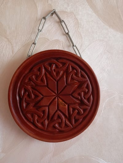 An ornate, circular wooden carving with intricate knotwork, possibly a decorative plate or wall hanging, is suspended by a metal chain against a textured, light-colored wallpaper. The carving appears to be a rich, dark reddish-brown wood with a polished finish, showcasing a star-like pattern in its center that expands outwards into interlacing Celtic knots. The wallpaper has a subtle, raised pattern reminiscent of flowing fabric or feathers, creating a soft, diffused background. The chain is simple, made of rectangular metal links, and attaches to the top of the carving, suggesting it is hung on a wall. The overall impression is one of decorative craftwork displayed in an indoor setting, likely a domestic environment. The specific location in Kyshtym, Russia, is not discernible from the image itself.