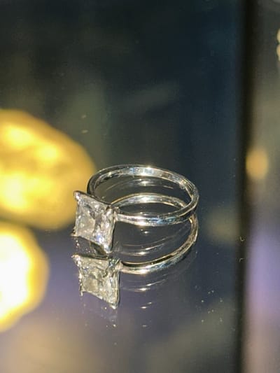 The image displays a close-up view of a diamond ring with a square-cut center stone, resting on a reflective surface. The ring is silver-toned and appears to be a single band. The background is dark and out of focus, with blurry yellow and purple lights visible, suggesting an indoor setting with artificial lighting. There is no indication of the city of Wittenberg, Germany, or any people present in the image.