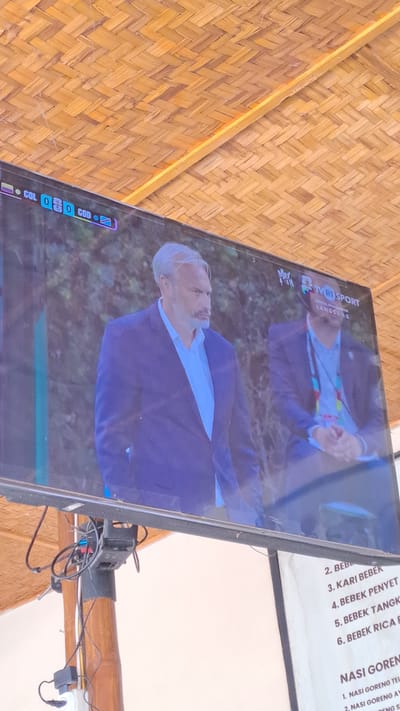 The image shows a television screen displaying a football match. The score displayed at the top left of the screen indicates COL 0 - 0 COD. The TV is mounted under a woven bamboo ceiling. Below the TV screen, a menu is partially visible, listing food items such as "BEBEK" (duck) and "NASI GORENG" (fried rice). The menu is written in Indonesian. The location appears to be an outdoor setting, possibly a cafe or restaurant, in Julikeude Dua, Indonesia, given the context of the menu.