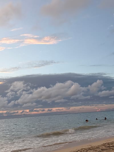 The image displays a beach scene in Roatán, Honduras, under dusk conditions. The sky transitions from a light blue at the zenith to a prominent horizontal band of dense, dark grey clouds mid-frame, with lighter grey cumulus clouds beneath. A subtle orange-pink luminescence is visible along the distant horizon. The ocean is a calm, light blue-grey, with small, white-foamed waves breaking at the shoreline. Three individuals are observed in the water, partially submerged with their heads and upper torsos visible. One individual is situated towards the left-center, while two others are grouped together further to the right. The immediate foreground consists of a strip of light brown sand, appearing wet near the water's edge.