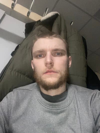 A man with light skin and a reddish-brown beard poses for a selfie. He wears a gray sweatshirt with a dark gray crew neck. Behind him lies an olive green jacket. Part of a metal railing and a beige wall are visible in the background, suggesting an indoor setting. His blue eyes are open and looking directly at the camera. The picture is shot at an angle.