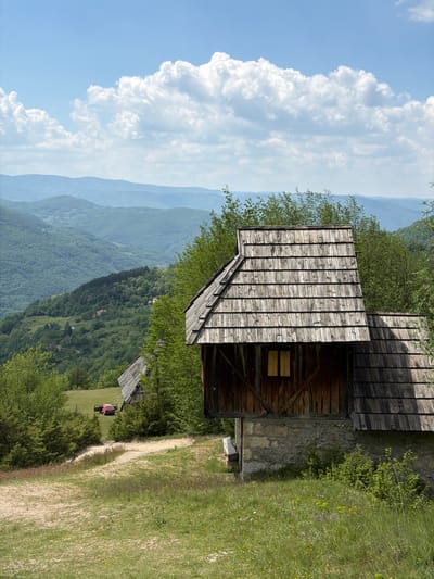 A landscape photo taken in the Prijepolje Municipality, Serbia, shows an overcast sky with tall cumulus clouds on the left, transitioning to a mountainous vista dotted with thick green forests.

To the right of the frame, a rustic, wooden structure with a planked roof and a weathered wooden wall rests atop a stone foundation, partially obscured by foliage. A dirt path meanders adjacent to this structure.

Further down the path and nestled within the greenery, there is a person seated at a small table with a red umbrella or cover.

The overall setting is one of a mountainous rural area, with nature and rustic construction harmoniously intermingling.