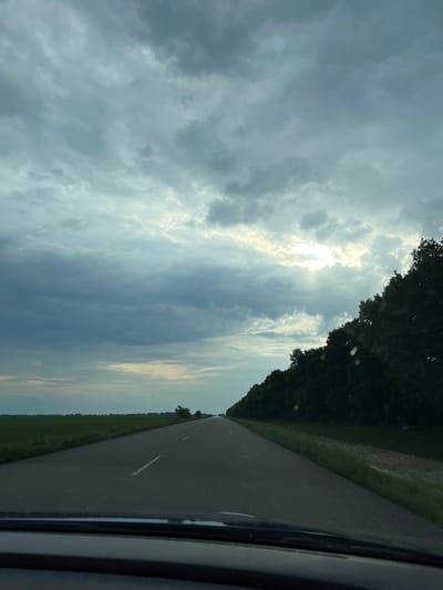 The image presents a forward-facing view from inside a moving vehicle on a straight, two-lane asphalt road in Petrivske, Ukraine. The road features a single dashed white line marking the center. To the left, a vast, flat expanse of green agricultural fields extends to the horizon. To the right, a dense, dark green treeline or forest borders the road and recedes into the distance. The sky above is predominantly overcast with layered, medium-grey clouds, showing lighter blue-grey patches. In the upper right quadrant of the sky, a prominent breakthrough in the cloud cover allows bright sun rays to penetrate. The bottom edge of the image reveals the top portion of a vehicle's dashboard or windshield frame. There are no individuals, other vehicles, or specific actions visible in the scene.