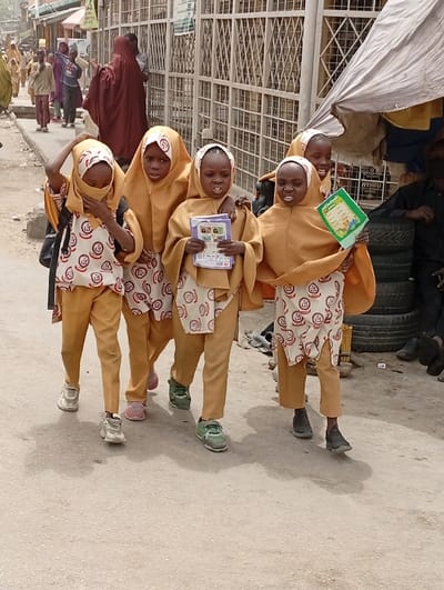 The image depicts five young girls in the Kofar Mazugal area of Nigeria, walking left-to-right on a paved street. All girls wear matching mustard-yellow full-length hijabs and trousers, paired with short-sleeved tunics featuring a red circular pattern on a white background. From left to right:
1. A girl with a black backpack partially visible, wearing white sneakers, holds her hijab over her lower face, exposing only her eyes.
2. A girl with pink slip-on shoes walks beside the first, looking forward.
3. A girl with green and white athletic shoes holds a small book with a colorful cover in both hands. Her expression suggests she is speaking or reacting.
4. A girl with black slip-on shoes holds a green book with both hands. Her mouth is open.
5. Partially visible, a fifth girl's face with a smiling expression is seen behind the fourth girl.
In the background, other individuals are visible walking on a sidewalk and further down the street. A woman in a dark red abaya walks away from the viewer. To the right, part of a market stall is visible with a light-colored tarp awning and stacked black tires. A metal security gate or fence separates the street from a building. The street appears dry and dusty.
