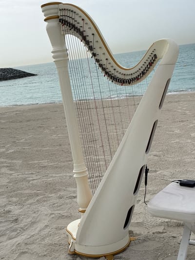A white and gold concert harp sits on a sandy beach, likely at Ghadeer Al Tayr, United Arab Emirates, based on the provided location. The harp's soundbox is angled towards the lower left of the image. The harp's strings stretch towards the upper right. A white chair and microphone are positioned near the soundbox. The backdrop includes beach sand and the ocean. The sky is overcast.