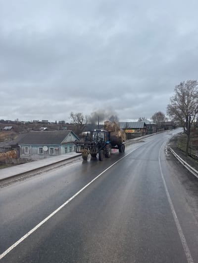 The image captures a rural or semi-rural road scene under an overcast, gray sky, suggesting a cool or cold day. The asphalt road, wet from recent precipitation, stretches into the distance, marked by a continuous white line along its edge.
In the foreground, a blue tractor with a front-end loader is parked or moving slowly on the right side of the road. Dark smoke billows from its exhaust pipe, indicating it is active. Behind the tractor, a trailer loaded with large, rounded hay bales is visible.
Along the left side of the road, a line of traditional wooden houses with pitched roofs can be seen. One prominent house is painted light blue with a darker gray roof and features multiple windows and a satellite dish on its facade. Bare trees, stripped of their leaves, dot the landscape, suggesting late autumn, winter, or early spring. A yellow and red chevron sign, typically indicating a sharp curve or hazard, is visible further down the road, past the tractor. The overall setting points to a small town or village road in Богородск, Russia. No specific people are visible.