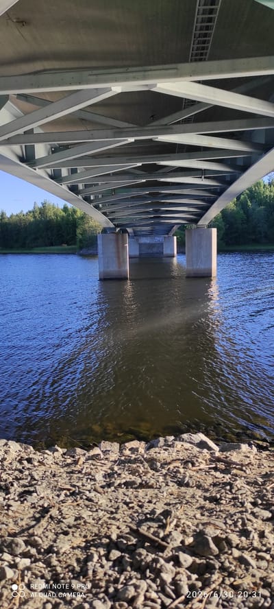 The image, taken in Hartola, Finland on June 30, 2026, at 20:31, captures a low-angle view from beneath a bridge. The bridge's concrete pillars rise from the water, supporting a complex network of metal beams. The water, reflecting the sky and surrounding trees, ripples gently. The foreground is a rocky shoreline with scattered debris. The scene depicts a natural environment integrated with man-made infrastructure.