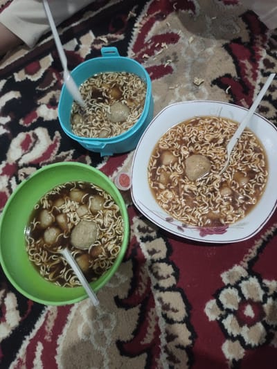 Three bowls of instant noodles with meatballs are arranged on a patterned carpet. A person's arm and shoulder, wearing a light-colored garment, are visible in the upper left corner, holding a white plastic utensil in the blue bowl. The blue bowl is filled with noodles and broth, containing several meatballs. To the right, a white bowl with a floral pattern also contains instant noodles and meatballs, with a white plastic utensil inside. To the left, a green bowl, similar to the blue one, is filled with noodles, broth, and meatballs, with a white plastic utensil submerged. A small red condiment dish is placed between the blue and white bowls. The carpet has a red and beige pattern. The scene appears to be an indoor setting, likely a home, given the carpet and casual arrangement of the food. There is no specific indication of the city of Cintadamai, Indonesia, in the image itself.