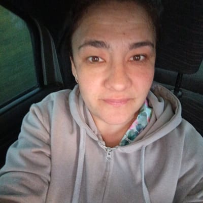 This is a selfie of a woman inside a vehicle. The woman has brown eyes and her hair is pulled back. She is wearing a light-colored hooded sweatshirt with a zipper. The background shows part of the vehicle's interior, including a seat and a window with a blurred green landscape visible outside. The image appears to have been taken during the day, likely with ambient lighting. The woman's expression is neutral. There is no visible text in the image. The scene suggests a casual moment, possibly while traveling.