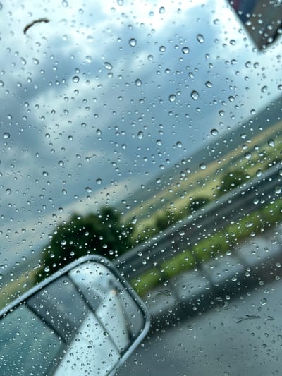 The image depicts a close-up view of raindrops on a car windshield. The foreground is dominated by numerous water droplets, varying in size, distorting the view of the scene beyond. The background shows a blurred, diagonal impression of an outdoor environment, suggesting a road or pathway with green foliage. A silver-colored car mirror is visible in the lower right portion of the frame, also partially obscured by raindrops. The sky appears cloudy and gray, indicating overcast weather. The overall impression is that of a rainy day, possibly in the morning or afternoon given the ambient light. No people are visible in the image. There is no discernible text. The location context is Vakarel, Bulgaria.