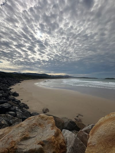 A wide-angle shot captures a beach scene under a cloudy sky in Coffs Harbour, Australia. The foreground is dominated by large, rugged rocks in varying shades of grey and brown, some with reddish-brown staining. To the left, a dark, craggy rock wall extends along the coastline.
Beyond the rocks, a wide, sandy beach stretches into the distance. The sand is a light beige color, marked with subtle footprints and a few scattered small stones. Gentle waves with white foam break on the shore, creating a wet sand area near the water's edge. The ocean water is a muted grey-blue, reflecting the overcast sky.
In the background, a low-lying, tree-covered coastline forms a distant horizon. Rolling hills covered in lush green vegetation are visible, with hints of buildings and structures in the far distance. The sky is filled with a dense, textured pattern of grey clouds, with patches of brighter sky and sunlight peeking through in places, creating a dramatic, diffused light. There are no visible people or animals in the frame.