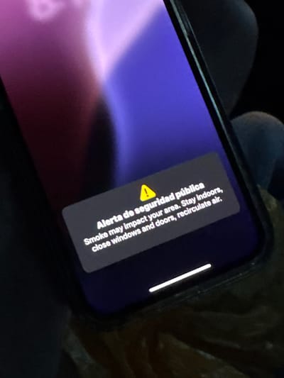 The image shows a smartphone displaying a public safety alert related to smoke. The alert is written in both English and Spanish, advising people to stay indoors, close windows and doors, and recirculate air, indicating a potential environmental hazard. The phone is positioned at an angle, and part of its screen displays a purple and pink gradient background. The setting is nondescript, with dark, blurred textures in the background. There is no specific information about the location within Hialeah, United States of America, nor are there any visible people or interactions in the image.