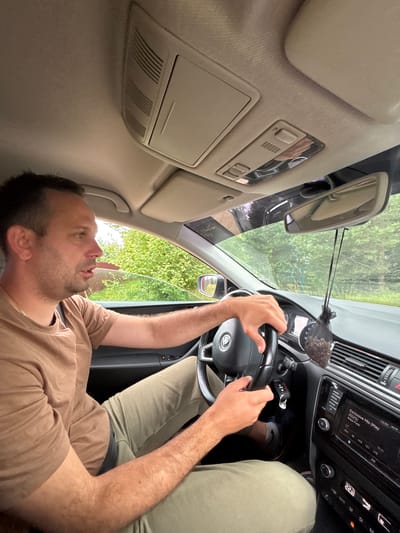 The image shows a man driving a car. He is wearing a brown t-shirt and light green pants. The car's interior is visible, including the steering wheel, dashboard, and rearview mirror. Outside the car, green trees and foliage can be seen, suggesting a rural or suburban environment. The image was taken in Anteplevo (Okhotinskoe S/P), Russia.