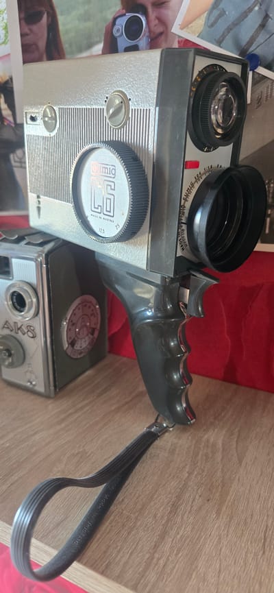 The image displays a collection of vintage cameras, primarily a silver and black Eumig film camera with a prominent lens and handle. To the left, a smaller, silver camera with "AK8" inscribed on its side is visible. The cameras are arranged on a wooden surface, with a red backdrop behind them. The Eumig camera has a ribbed grip and a strap attached to its handle. The overall scene suggests a display or collection of photographic equipment. There is no indication of the city of Gostagayevskaya, Russia, in the image.