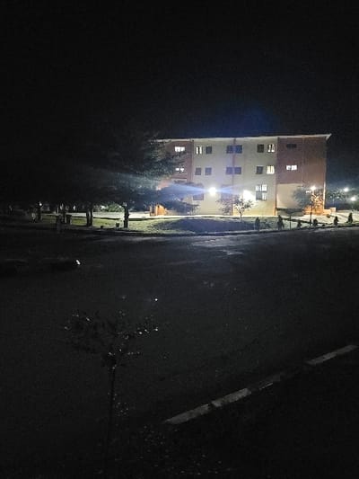 The image depicts a nighttime scene in Gaba, Nigeria, focusing on a four-story building. The building, constructed with a light-colored facade and reddish-brown vertical accents, is illuminated by numerous interior window lights and exterior ground-level fixtures. Several trees with dark foliage are positioned in the midground, between the building and the foreground. The foreground features a dark asphalt surface, possibly a road or parking lot, which exhibits reflective patches consistent with wetness. A concrete curb is visible along the lower right edge of the frame. No individuals, animals, or vehicles are present. The ambient lighting is exclusively artificial, originating from the building and other distant light sources, against a dark sky.
