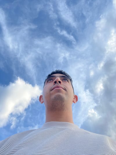 A low-angle, upward-facing photograph depicts a person with medium skin tone, short dark curly hair, and stubble, wearing a light-colored ribbed shirt. The person's face is angled downwards towards the camera. The background consists of a cloudy sky with varying shades of blue and white. The location and city are not discernible from the provided image. No specific actions or interactions are evident.
