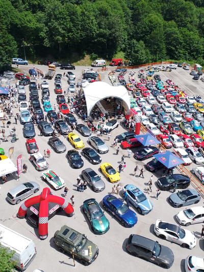 An aerial, high-angle, wide shot captures a car show or event taking place in a paved outdoor area surrounded by dense green trees. The scene is packed with a diverse array of vehicles, predominantly cars, parked in rows. Many of these appear to be performance or sports cars, with some vintage models and a few utility vehicles also present.
In the center of the frame, a large white tent structure with an open side is set up, likely serving as a central hub for the event. Red and white checkered inflatable arches stand in front of the tent entrance. Several tents and temporary structures, including two blue Red Bull branded umbrellas, are scattered throughout the area.
A significant number of people are visible, milling about the vehicles, observing the displays, and interacting with each other. Some individuals are standing near cars, while others are walking through the parking area. Event staff or organizers might be present, indicated by some people wearing vests or seemingly directing activities.
The environment suggests a location in a somewhat rural or mountainous area, given the dense tree line in the background and the paved, open space suitable for such an event. The presence of a white van with "Sara" branding suggests a possible sponsor or service provider. The overall impression is one of a busy, outdoor automotive gathering.
Specifically, the event is taking place at the Autodromo Internazionale di Rieti, located near the city of Rieti, Italy. The distinctive features of the track's layout and surrounding landscape, including the trees and hills, are consistent with this location.