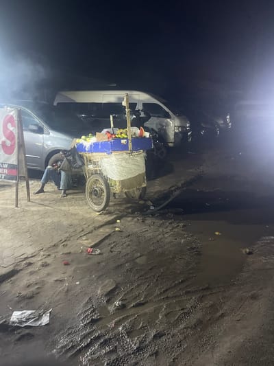 A street scene in Obiaruku, Nigeria at night. A man sits on a low seat beside a fruit cart filled with produce. The cart stands next to a light-colored van. The ground is uneven and muddy in places, strewn with litter. A sign is visible in the foreground, partly obscured by smoke or fog. The scene is dimly lit.