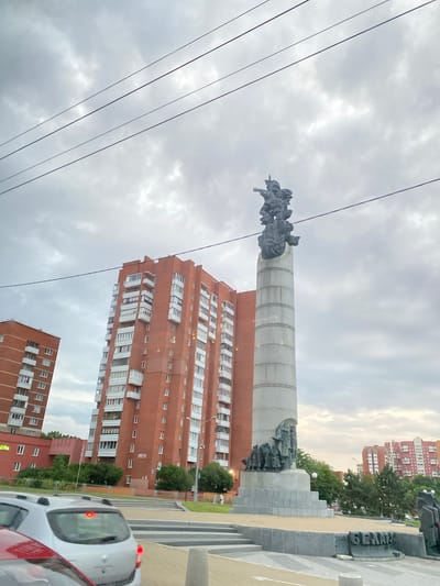 The image depicts a monument with figures on top of a tall, cylindrical structure. The monument is situated in an urban setting, with a tall, red brick apartment building visible behind it. In the foreground, there are power lines, and a white car is partially visible. The scene appears to be outdoors, possibly in a public square or park, given the presence of the monument. The sky is overcast, suggesting it could be daytime with cloudy weather. The overall atmosphere is somewhat solemn, likely due to the nature of monuments which often commemorate historical events or figures. The text "ЦВЕТЫ" (flowers) is visible on a sign on the building. The location is identified as Лугаваслабадскі сельскі Савет, Belarus.