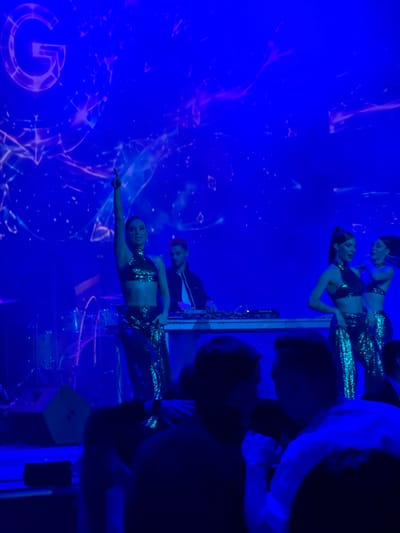 In Akhalubani, Georgia, a vibrant nightclub scene unfolds under intense blue lighting. A large, stylized "G" is projected onto a screen, serving as a backdrop for a performance. A female dancer, clad in a shimmering, sequined crop top and skirt, strikes a pose with her arm raised. To her right, a male DJ stands behind a console of equipment, appearing focused on his work. Further to the right, another female dancer, also in a sequined outfit, is captured mid-motion. In the foreground, blurred figures of audience members are visible, suggesting a lively atmosphere. The stage is further equipped with drums and speakers, indicating a live music or DJ set. The overall ambiance is energetic and celebratory, characteristic of a nightclub setting.