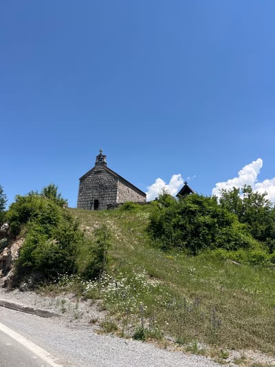 The image captures a stone church situated atop a grassy hill under a clear blue sky with scattered white clouds. The church, constructed of rough-hewn stones, features a bell tower topped with a cross and a small arched doorway. Green trees and bushes surround the church and the hill. In the foreground, a gravel and asphalt road with a white stripe runs along the bottom left of the frame. Wildflowers, predominantly white daisies and some purple flowers, are scattered across the grassy slope. The scene appears to be a tranquil rural landscape, possibly in or near Bakovići, Montenegro, given the context provided.