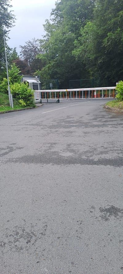 This is a medium shot, eye-level, outdoor, daylight, full frame photograph of the entrance to Breaffy House Resort in Breaffy, Ireland. The scene is a paved road leading to a security barrier. The barrier is a white, horizontal bar with orange stripes, currently lowered and blocking the entrance. To the left of the barrier is a small, gray security hut with a white awning and a sign that reads "BREAFFY HOUSE WELCOME CENTRE". A tall, gray metal pole with a security camera attached stands to the left of the security hut. Lush green trees and foliage line the sides of the road and surround the entrance, with a gray, overcast sky visible above the tree line. The asphalt road surface is wet and appears to have some dark patches, possibly from rain. No people are visible in the frame.
