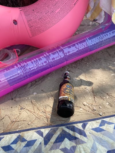 The image shows a scene on a sandy beach in Crikvenica, Croatia. A partially inflated pink pool float and a purple inflatable tube lie on the sand next to a patterned blue and white beach towel. A dark glass bottle with a swing-top cap, labeled "Hacker-Pschorr Münchner Gold," is also placed on the sand. The bottle appears to be a beer. Pine needles are scattered across the sand, suggesting an outdoor setting with some vegetation nearby. The lighting suggests it is daytime, possibly late afternoon, due to the long shadow cast by an unseen object or person. The overall mood is relaxed and leisurely, indicative of a beach outing. The text on the pink pool float is a warning label in multiple languages, detailing safety instructions for the product.