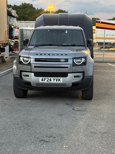 A silver Land Rover Defender is parked on a grey asphalt surface. The vehicle is facing the camera, and its front license plate reads "AF24 YVK." The Defender has its headlights on, emitting a bright white light. Behind the Defender, a dark grey, box-like trailer is attached. To the left of the Defender, a portion of a black truck with red and white detailing is visible. To the right, a white trailer with horizontal red and yellow stripes is parked, displaying the text "www.groupe" on its side. The background consists of a metal fence, trees, and a cloudy sky with visible streetlights. The scene appears to be an outdoor parking area, possibly a rest stop or service area. There are no people visible in the image. The location is identified as Grenay, France.