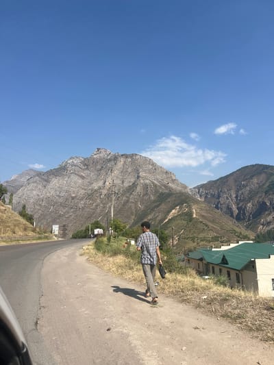 A man walks on a dirt path beside a road in a mountainous region of Yakkatut, Uzbekistan. The man is wearing a plaid shirt and grey pants. He is carrying a black bag in his right hand. In the background, there are steep, rocky mountains and a clear blue sky with a few white clouds. To the right of the road, there are some buildings with green roofs. The road is paved and curves to the left. The area is dry and grassy, with scattered trees. Power lines run along the left side of the road.