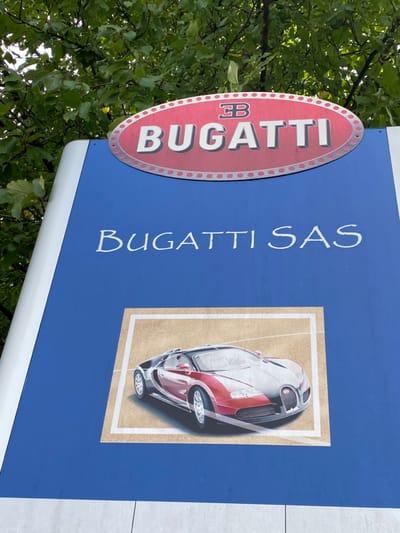 A large, vertical blue sign for Bugatti SAS is situated outdoors in Dorlisheim, France, partially obscured by green tree foliage in the upper background. The sign's upper portion features a red, oval-shaped emblem with a silver-dotted border, displaying "BUGATTI" in white capital letters and a stylized "EB" monogram above it. Directly below the emblem, "Bugatti SAS" is written in white, cursive-style script. The lower section of the sign contains a rectangular graphic of a two-tone Bugatti Veyron 16.4 supercar, depicted with a red front-left portion and a dark gray/black rear-right portion. This graphic is set against a light brown background with a white border. The sign itself has a white vertical border on its left side. No individuals or specific actions are visible in the scene.