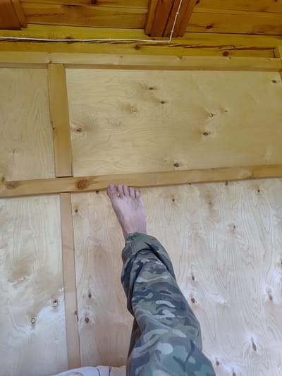 In a wooden room, a barefoot person wearing camouflage pants is shown from the waist down, with their left leg extended upwards against a plywood wall. The room's walls and ceiling are constructed from light-colored wood planks and plywood. A white cable is visible running across the ceiling. The environment suggests an indoor, likely unfinished, space. The specific location of Бетюнцы, Russia, is not discernible from the visual information presented in the image.