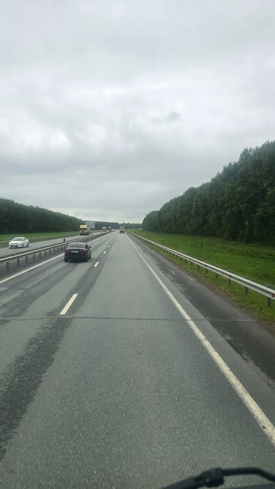 The image depicts a highway in Staroye Baysarovo, Russia, on a cloudy day. The wet asphalt suggests recent or ongoing rain, with reflections visible on the road surface. The perspective is from a vehicle traveling on the highway, looking forward and slightly to the right.
The highway has multiple lanes, separated by white lines. A metal guardrail runs along the right side of the road, separating it from a grassy verge and a dense line of green trees. In the distance, several vehicles are visible, including a yellow truck, a black car, and a white car in the foreground. The sky is overcast and gray, creating a muted lighting condition.
The scene appears to be a daytime view, given the visibility of the road and surrounding landscape. No people are clearly discernible, though the presence of vehicles implies human activity. The overall mood conveyed by the image is one of travel on a somewhat somber, overcast day, with the wet road and trees suggesting a natural, possibly rural, setting. No discernible text is visible in the image.