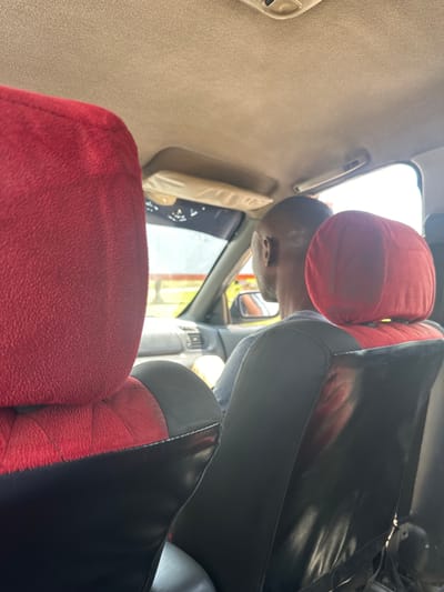 An interior view from the back seat of a car shows a bald Black man driving. The car's interior has red and black seat covers. The man is wearing a grey shirt. Through the windshield, a yellow and red object is visible, possibly a building or a sign. The overall scene suggests an ordinary moment of travel in Wupa, Nigeria.