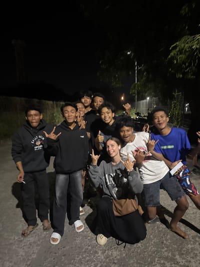 A group of ten young people, predominantly male, pose for a photograph outdoors at night. The scene appears to be in a semi-urban or rural area, possibly near Labuhan Lombok, Indonesia, as indicated by the presence of trees, a dirt or gravel pathway, and faint distant lights suggesting a town or industrial area. The lighting is dim, with artificial light sources casting shadows.

In the foreground, a young woman with light-colored hair is crouched down, wearing a grey hoodie and dark pants. She is smiling and making a hand gesture. To her right, a young man in a white t-shirt and dark shorts is standing, also smiling and making a hand gesture. To his right, another young man in a blue t-shirt is visible.

Behind them, a group of young men are arranged, some smiling and making various hand gestures. They are dressed casually in t-shirts and hoodies. One of the men on the far left of the frame appears to be holding a cigarette. The background includes trees, a tall structure resembling a light pole or tower in the distance, and what looks like a motorcycle parked further back. The overall mood seems to be casual and celebratory.