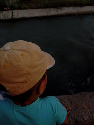 A young child wearing a light brown baseball cap and a teal t-shirt is seen from behind, looking at a dark body of water. The child's right ear is visible. The water appears to be in a canal or a similar man-made waterway, bordered by a concrete structure. The background shows some vegetation along the far bank of the waterway. The image is dark, suggesting it was taken in low light conditions, possibly during dusk or in a shaded area. The setting is identified as Kızıl-Bel, Kyrgyzstan.