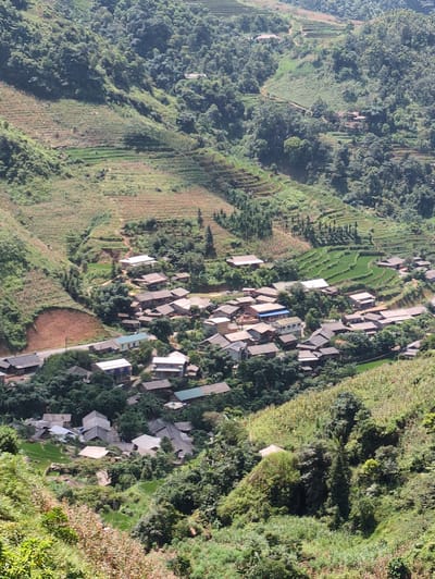 This image shows a high-angle, vertical view of a rural village nestled in a valley in Minh Son Commune, Vietnam. The landscape is characterized by lush green, terraced rice paddies on the steep hillsides, interspersed with forested areas. A dirt road winds through the village, which consists of numerous houses with dark-tiled roofs, many of them clustered together. The houses appear to be made of wood and brick. The environment is predominantly natural, with dense vegetation covering most of the terrain. There are no visible people in the image. The time of day appears to be daytime, with bright sunlight casting shadows.
