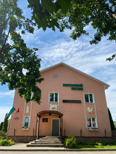 The image presents a bright daylight scene featuring a two-story, peach-colored building with a gable roof, set against a blue sky with scattered white clouds. Lush green trees frame the composition on the left and bottom.
The building's facade prominently displays "ЛЮБАНСКИЙ ЛЕСХОЗ" (LYUBANSKY LESKHOZ) in vertical green letters, indicating it is likely the Lyuban Forestry or Timber Enterprise. Below this text is a dark rectangular digital display showing illuminated numbers, possibly functioning as a clock or thermometer. Several white-framed windows punctuate the walls. Decorative white relief sculptures of animal heads, resembling deer or similar horned creatures, are mounted on the wall, with one positioned above the main entrance. The entrance features a brown canopy and steps leading up to a dark door.
To the lower left of the entrance, two flags, one green and one red, are visible. A street sign below the flags clearly reads "ПЕРВОМАЙСКАЯ" (PERVOMAYSKAYA), translating to "First of May Street," followed by the number "59." The base of the building is adorned with green bushes and shrubs, and a paved sidewalk runs along the right side of the frame.
The image captures a calm, static scene under clear, sunny weather, suggesting it was taken during midday or early afternoon. No people or specific activities are depicted. The location is Lyuban, Belarus, as indicated by the context and the readable text on the building and street sign.