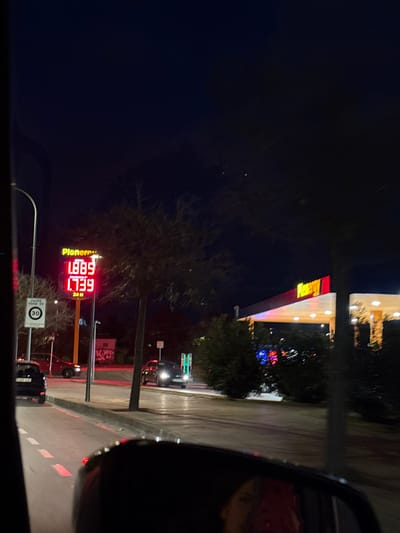 The image displays a nighttime urban scene in Les Meravelles, Palma de Mallorca, Spain, observed from within a vehicle. On the left, a tall, illuminated digital sign for "Pionero" gas station displays fuel prices: "1.889" and "1.739" (likely representing Euros per liter for different fuel types, with "GASOLINA 95" explicitly visible for the latter), and "24H" indicating continuous operation. Below the sign, a circular traffic sign indicates a "30" km/h speed limit, with the word "PALMA" partially visible beneath it. To the right, a brightly illuminated "Energy" brand gas station canopy, featuring yellow and red signage, extends over several fuel pumps. Multiple dark-colored vehicles are present on the paved road and within the gas station forecourt, some with active headlights. Bare deciduous trees are interspersed throughout the scene, alongside green shrubs near the gas station property. A distorted reflection of a human face is visible in the vehicle's rearview mirror at the bottom center. The sky is dark, with artificial lighting providing primary illumination.