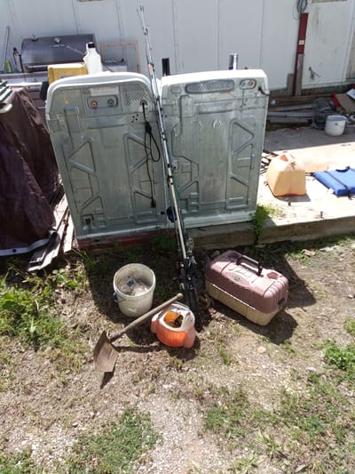 This outdoor scene in Park Hill, United States shows a collection of discarded or stored items against the backdrop of a light-colored, possibly metal-sided building. Two upside-down, identical appliances, likely washing machines, dominate the center of the frame. A pair of fishing rods leans against one of the appliances. To the left, a covered object sits on a stack of planks. To the right are containers and materials in a messy pile. In the foreground, there is a shovel lying on the ground next to a white bucket, an orange container, and a closed, pink-and-beige fishing tackle box. The ground is a mix of dirt, gravel, and sparse vegetation.