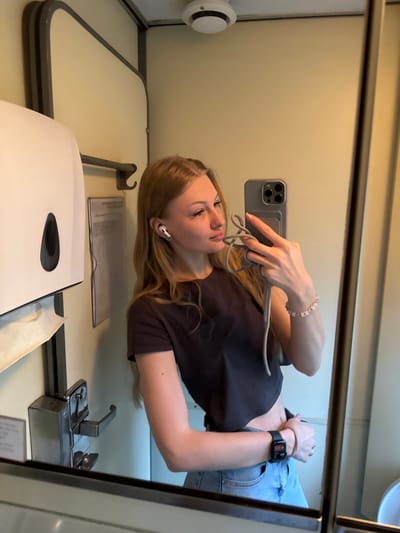 A young Caucasian woman with long blonde hair is taking a mirror selfie inside a train compartment. She is wearing a dark t-shirt, light blue jeans, and white earbuds. She has a bracelet on her left wrist and a smartwatch on her right wrist. She is holding a gray smartphone in her right hand. The train compartment appears to be a bathroom, with a white paper towel dispenser on the left wall, a metal faucet below it, and a mirror reflecting the scene. The walls are light yellow. There is a smoke detector on the ceiling. The overall impression is of a casual selfie taken in a small, utilitarian space. The location is described as Nizhnyaya Baskaya, Russia.