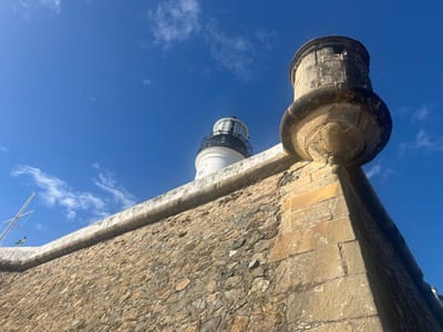 The image displays a low-angle perspective of the Forte de Santo Antônio da Barra and the Farol da Barra (Barra Lighthouse) in Salvador, Brazil, against a clear blue sky. A large, textured stone fort wall, constructed from irregular brown, grey, and ochre-colored blocks, dominates the foreground, extending from the lower left to the right. A lighter-colored, smooth stone coping runs horizontally along the wall's top edge. On the right, a cylindrical, weathered stone bartizan (watchtower) with small rectangular openings protrudes from a corner of the fort wall, featuring a decorative base. Behind the fort wall, to the left of the bartizan, the upper portion of the white Farol da Barra lighthouse is visible, capped by a black lantern room. To the far left, a bare mast, likely from a vessel or a flagpole, rises above the wall, from which a small green and yellow flag flutters. The background consists of a vibrant blue sky with sparse, wispy white clouds. The scene is brightly illuminated by natural sunlight, casting distinct shadows and emphasizing the textures of the stone structures. No people are present.