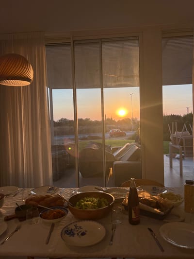 An interior view captures a set dining table near a window. Outside, a vibrant sunset paints the sky. The window frame divides the outdoor scene into layers of a suburban street and a green space. The table setting includes plates, salad, bread, drinks and other unknown dishes. A decorative lamp is visible near the window, adding an extra layer of warmth to the scene.