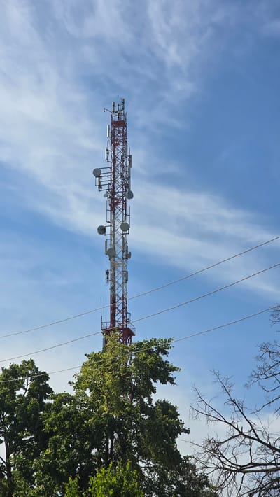 The image shows a tall telecommunications tower against a bright blue sky with wispy clouds. The tower is red and white and is adorned with multiple antennas and dishes. In the foreground, lush green trees partially obscure the base of the tower, and bare branches from another tree are visible to the right. Power lines stretch horizontally across the frame, in front of the tower. The scene is outdoors, with the focus on the technological structure against a natural backdrop. The image does not depict any people or specific urban landmarks to indicate a precise location within the city of Актюбинский, Russia.