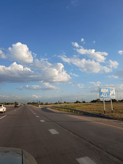 The image shows a highway on a sunny day with scattered clouds. A white car is visible on the left side of the road, driving in the same direction as the viewer. On the right side of the road, there is a blue road sign with white text and an arrow. The text on the sign is in Cyrillic and Latin alphabets, reading "АЛИ" and "ALI" respectively, with a white arrow pointing to the right. The highway is lined with guardrails and occasional streetlights. In the background, there are trees and power lines, and in the distance, mountains are visible. The scene appears to be in an arid or semi-arid region, with dry grass lining the side of the road. The overall impression is of a typical highway scene in a suburban or rural area.