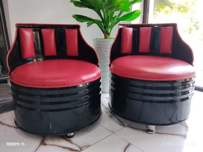 Two barrel-shaped chairs with red seats and backs are positioned on a tiled floor. The chairs have black exteriors with red cushioning in the back and seat. A potted plant with large green leaves sits between the chairs. The background appears to be a window with diffused sunlight. A timestamp "21.06.2026 10:56" and the text "REDMI 15" are visible in the lower right and left corners of the image respectively. There are no people visible in the image. The scene is indoors.