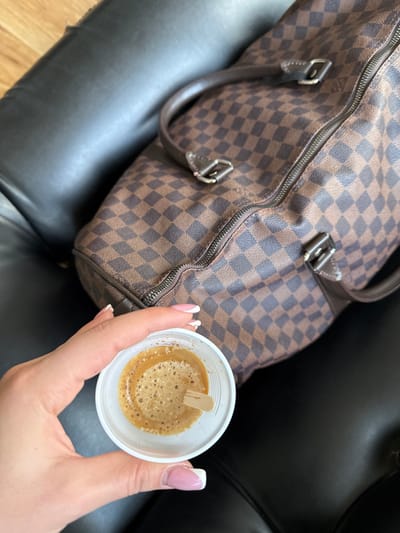 A close-up, high-angle shot features a left hand, displaying a French manicure, holding a small white disposable plastic cup. The cup contains a dark brown liquid topped with a light brown foam (crema) and a small wooden stirrer stick. In the background, a large brown and dark brown checkered patterned bag, consistent with the Louis Vuitton Damier Ebene design, rests on a black upholstered surface, likely a seat. The bag has visible brown trim, handles, and a top zipper. A portion of a light brown wooden surface is visible in the upper left background. The specific location in Senokos, Bulgaria, cannot be determined from the visual information presented.